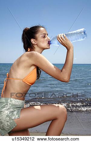 Stock Image - Athletic female young adult drinking bottled water on beach. Fotosearch