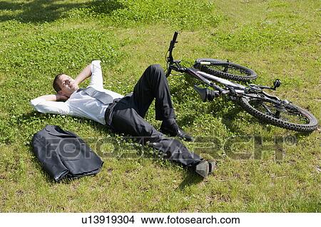 Businessman lying down in grass next to bicycle enjoying the sun View Large Photo Image Picture - Businessman lying down in grass next to bicycle enjoying the sun. Fotosearch