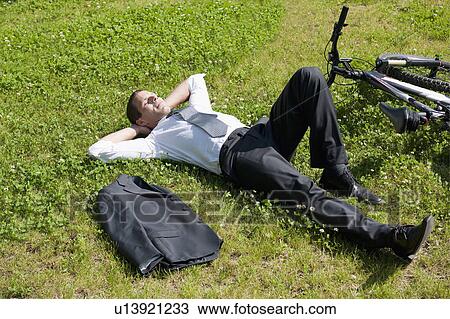 Stock Image - Businessman lying down in grass next to bicycle enjoying the sun. Fotosearch