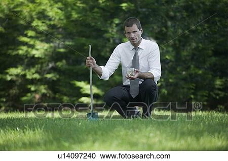 Stock Image - Businessman on lawn with shovel and jar of coins. Fotosearch