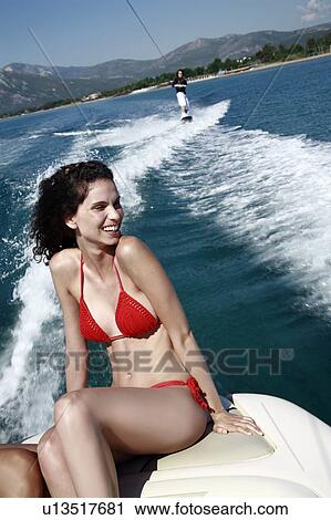 Stock Image - Female young adult on speedboat; male waterskiing. Fotosearch