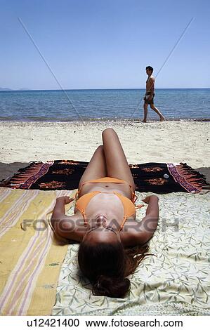 Stock Image - Female young adult sunbathing; man walking by. Fotosearch