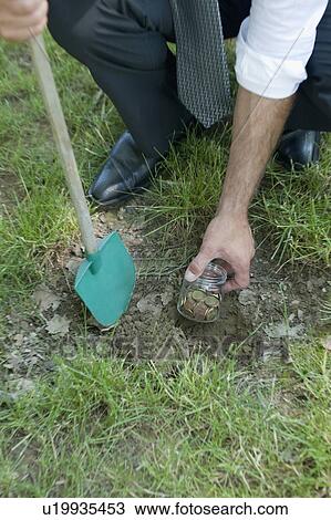 Stock Image - Hand with shovel hiding jar of coins in grass. Fotosearch