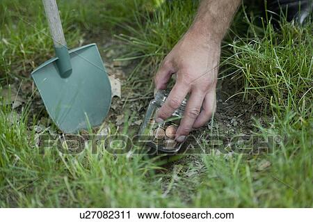 Stock Image - Hand with shovel hiding jar of coins in grass. Fotosearch