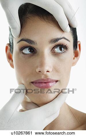 Stock Image - Hands wearing surgical gloves holding young woman's face. Fotosearch
