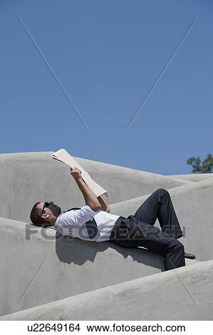 Picture - Man lying down on wall reading newspaper. Fotosearch