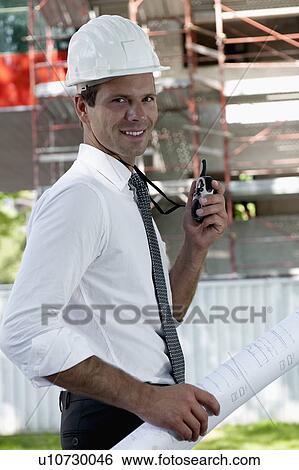 Stock Photograph - Man with hardhat, walky talky radio, and architectural plans at construction site. Fotosearch