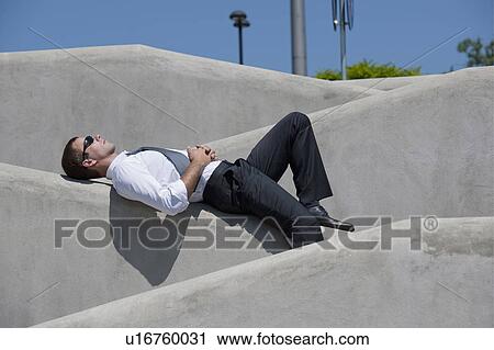 Stock Image - Man with tie and sunglasses lying down on wall. Fotosearch