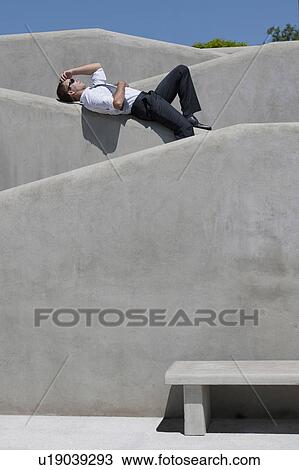Stock Image - Man with tie and sunglasses lying down on wall. Fotosearch