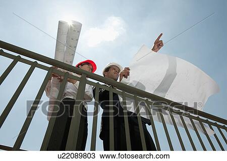 Stock Image - Professionals holding plans at construction site. Fotosearch