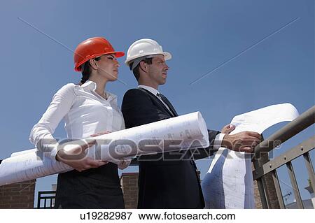 Professionals holding plans at construction site View Large Photo Image Stock Photo - Professionals holding plans at construction site. Fotosearch