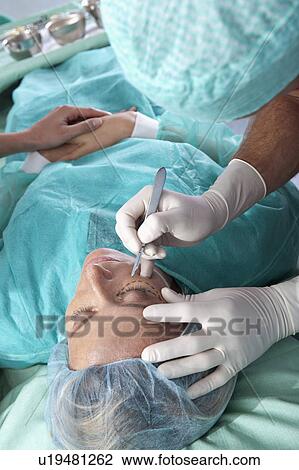 Stock Image - Surgeon with scalpel to patient's face. Fotosearch