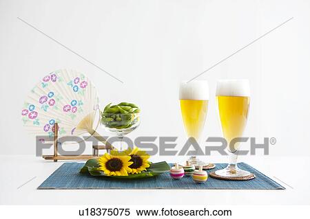 Stock Photography - Glasses of beer, edamame, sunflowers, tops and fan on place mat. Fotosearch