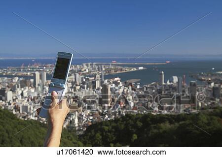 Stock Image - Man's hand holding a cell phone and cityscape of Kobe, Hyogo Prefecture, Honshu, Japan. Fotosearch