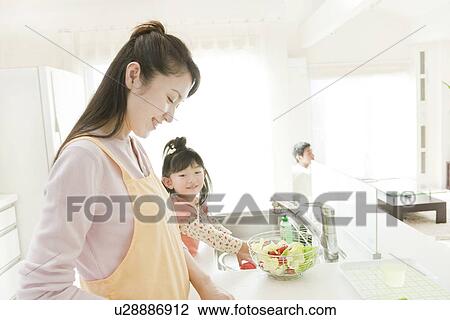 Stock Image - Mother and Daughter Cooking, Father in Background. Fotosearch