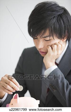 Stock Photo - Young businessman putting coin into piggy bank. Fotosearch