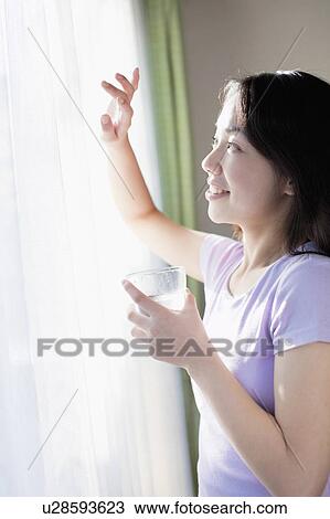 Stock Image - Young Woman Looking Through Window in Morning. Fotosearch