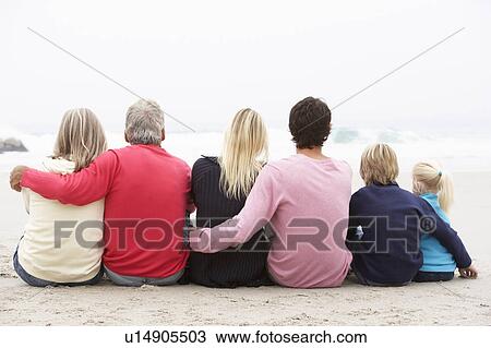 Stock Image - Back View Of Three Generation Family Sitting On Winter Beach Together. Fotosearch