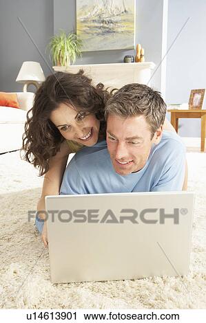 Stock Image - Couple Using Laptop Relaxing Laying On Rug At Home. Fotosearch
