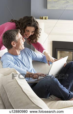 Stock Image - Couple Using Laptop Relaxing Sitting On Sofa At Home. Fotosearch