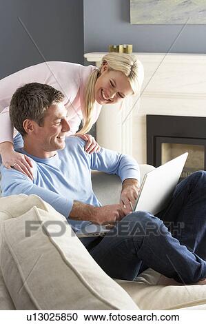 Stock Image - Couple Using Laptop Relaxing Sitting On Sofa At Home. Fotosearch
