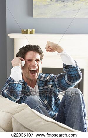 Stock Photo - Excited Man Sitting On Sofa At Home Talking On Phone. Fotosearch