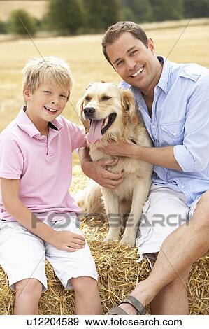 Stock Photo - Father And Son Sitting With Dog On Straw Bales In Harvested Field. Fotosearch