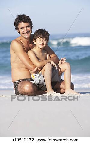 Stock Image - Father And Son Wearing Swimwear Sitting On Sandy Beach. Fotosearch