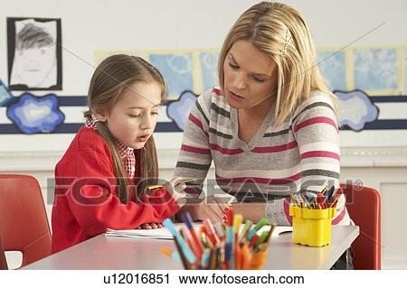 Stock Image - Female Primary School Pupil And Teacher Working At Desk In Classroom. Fotosearch