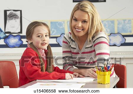 Stock Photo - Female Primary School Pupil And Teacher Working At Desk In Classroom. Fotosearch