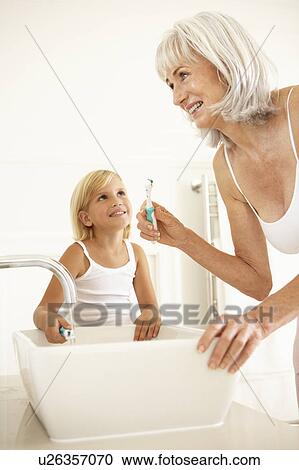 Stock Image - Grandmother Brushing Teeth In Bathroom With Granddaughter Watching. Fotosearch