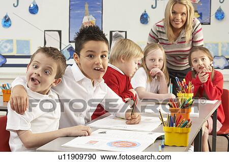 Stock Image - Group Of Primary Schoolchildren And Teacher Working At Desks In Classroom. Fotosearch
