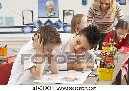 Stock Image - Group Of Primary Schoolchildren And Teacher Working At Desks In Classroom. Fotosearch