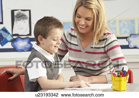 Stock Image - Male Primary School Pupil And Teacher Working At Desk In Classroom. Fotosearch