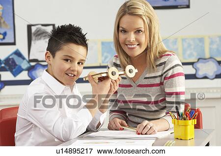 Stock Photo - Male Primary School Pupil And Teacher Working At Desk In Classroom. Fotosearch
