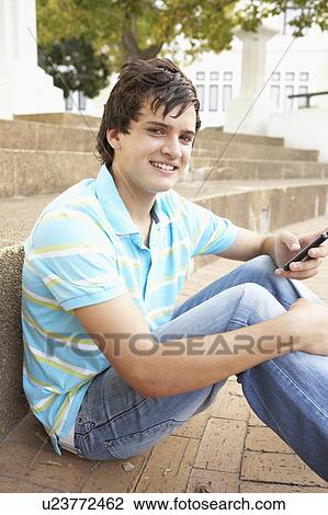 Stock Image - Male Teenage Student Sitting Outside On College Steps Using Mobile Phone. Fotosearch