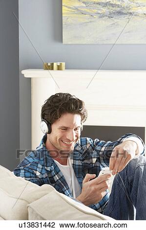 Stock Image - Man Listening To MP3 Player On Headphones Relaxing Sitting On Sofa At Home . Fotosearch