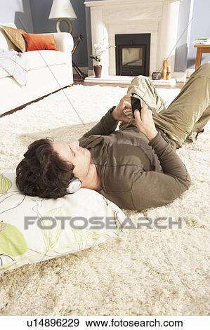 Stock Photo - Man Listening To MP3 Player On Headphones Relaxing Laying On Rug At Home . Fotosearch