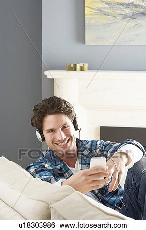 Stock Photograph - Man Listening To MP3 Player On Headphones Relaxing Sitting On Sofa At Home . Fotosearch