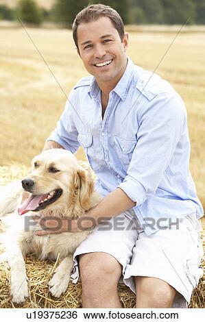 Stock Photograph - Man Sitting With Dog On Straw Bales In Harvested Field. Fotosearch