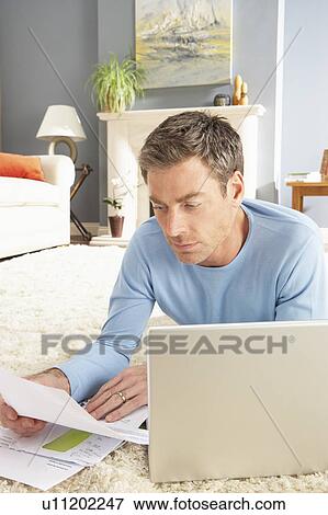 Stock Photo - Man Using Laptop To Manage Household Bills Laying On Rug At Home . Fotosearch