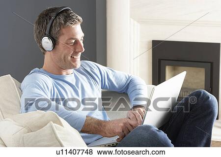 Stock Photo - Man Using Laptop Wearing Headphones Relaxing Sitting On Sofa At Home . Fotosearch