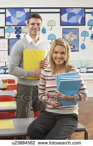 Stock Image - Portrait Of Male And Female Teacher Sitting At Desk In Classroom. Fotosearch