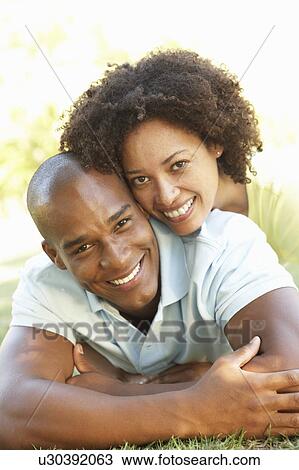 Stock Image - Portrait Of Young Couple Laying On Grass In Park. Fotosearch