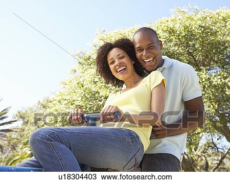 Stock Image - Portrait Of Young Couple Riding On SeeSaw In Park. Fotosearch