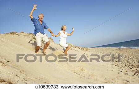 Stock Image - Senior Couple Enjoying Beach Holiday Running Down Dune. Fotosearch