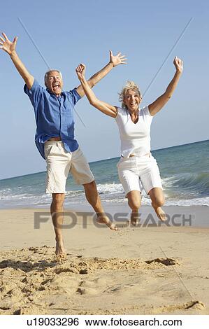 Stock Photograph - Senior Couple Enjoying Beach Holiday Jumping In Air. Fotosearch