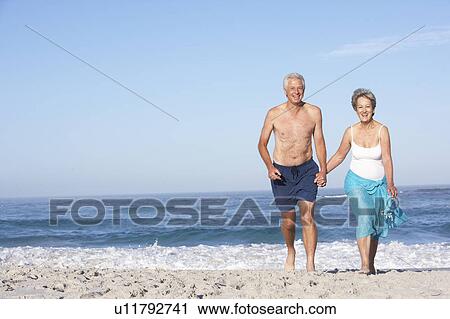 Stock Image - Senior Couple On Holiday Running Along Sandy Beach. Fotosearch
