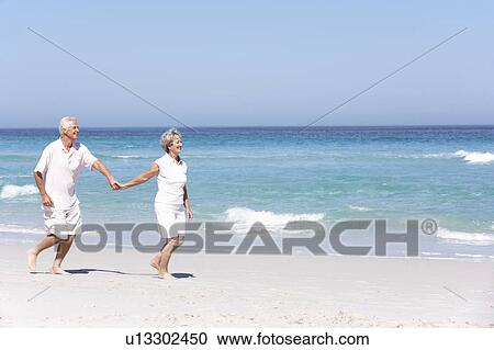 Stock Image - Senior Couple On Holiday Running Along Sandy Beach. Fotosearch