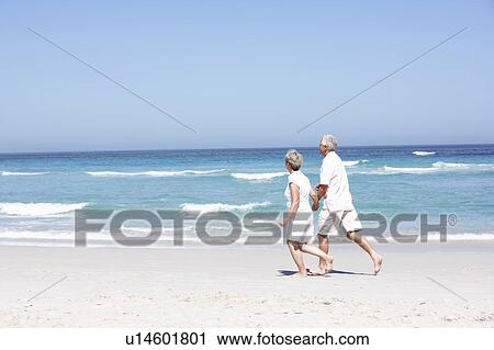 Stock Image - Senior Couple On Holiday Running Along Sandy Beach. Fotosearch